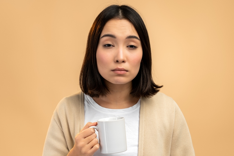 眠そうな表情でマグカップを持つ女性。むくみが出やすい朝のイメージ